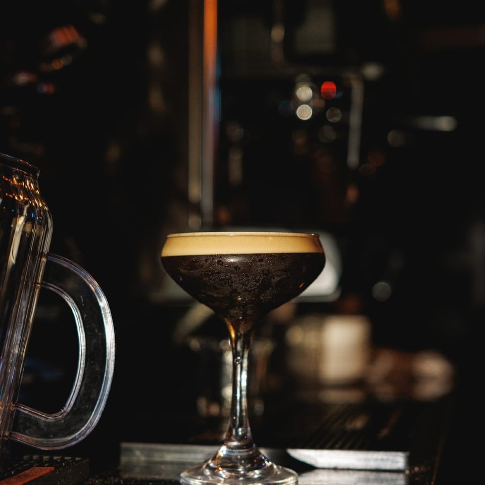 Espresso martini on the dark bar beside bar equipment.