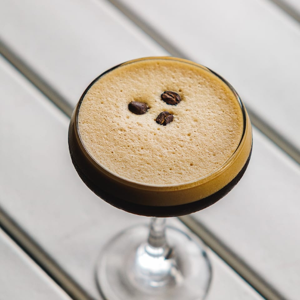 Espresso martini topped with three coffee beans against a bright slatted background.