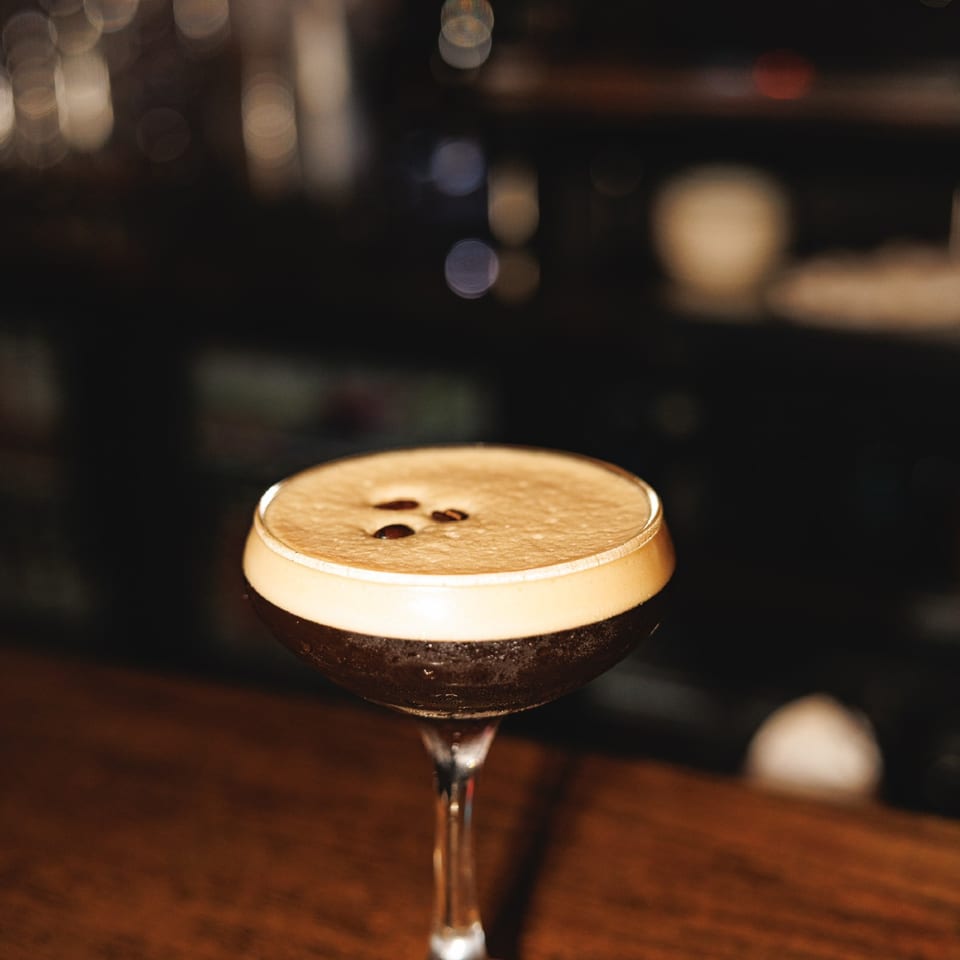 Foamy espresso martini on a wooden bar with warm background lights.